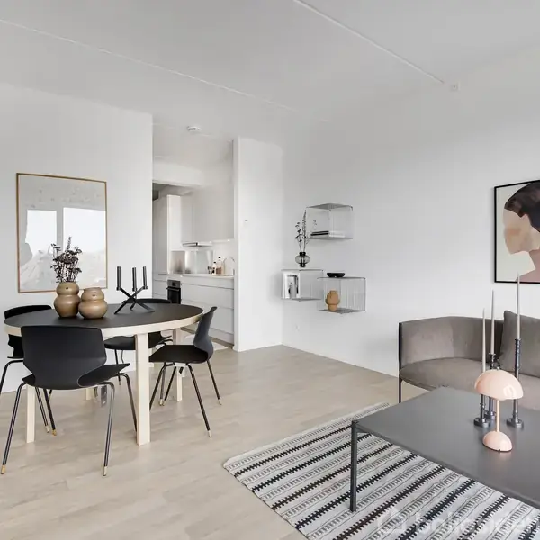Et moderne stue-spisestueområde med et rundt spisebord omgivet af sorte stole. En grå sofa står ved siden af et sofabord med en lyserød lampe. Kunst hænger på de hvide vægge.