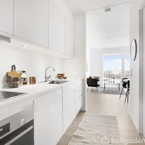 Hvidt køkken med moderne apparater, indbygget ovn og kogeplade, diskret indretning. I baggrunden ses lys stue med sofa, stol og adgang til balkon gennem en stor glasdør.