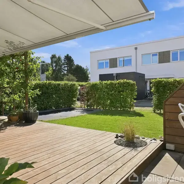 Hvidt, moderne to-etagers rækkehus står stille på en flisebelagt indkørsel, omkranset af grønt græs, med en sort carport foran. Blå himmel og naboer i baggrunden.