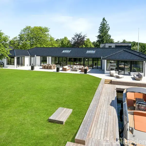 Et moderne, en-etagers hus med store vinduer og sort tag står blandt grønne træer. Foran huset er en velplejet græsplæne og en bred, trædækket terrasse ved en bådebro med en båd.