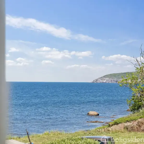 Hav møder kysten med klipper og grønt græs ved siden af. En bakke stiger langs højre kant under en klar blå himmel med lette skyer.