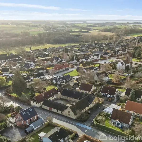 Et kvarter med spredt bebyggelse af huse ses fra luften. Husene er omgivet af træer og marker, hvilket skaber en landlig atmosfære. Ingen tekst fundet.