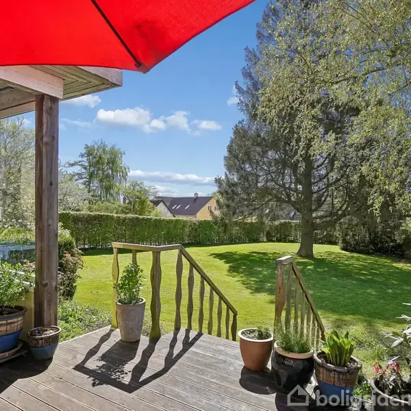 Verandaen har blomsterkrukker og en rød parasol. Den åbner ud til en velplejet græsplæne omgivet af høje træer og en hæk. Himlen er blå med få skyer.