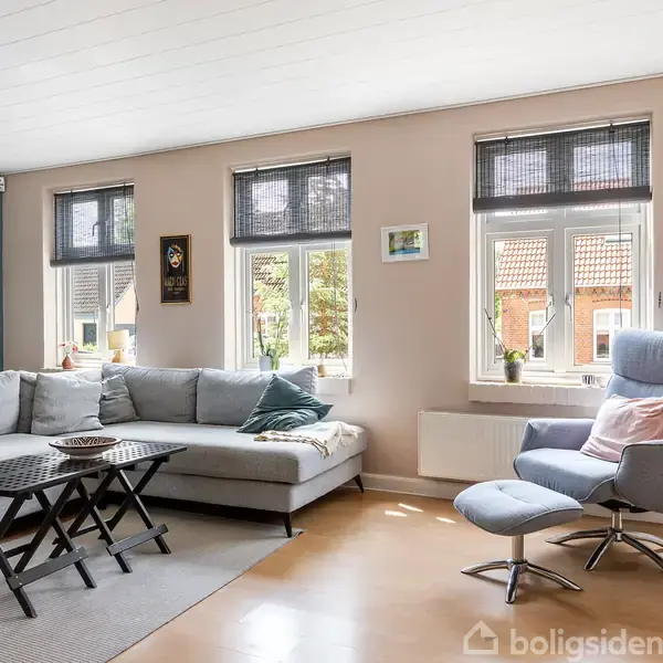 En lys stue med en stor, lysegrå hjørnesofa og et blåt lænestol-arrangement. Sofaen står ved siden af tre vinduer, der lukker lys ind. Væggene er dekoreret med billeder.