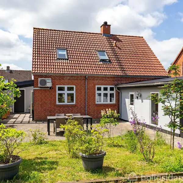 Et rødt murstenshus med skråt tag har en have i forgrunden. Havemøbler står på terrassen, mens græs og blomster omgiver det. Skyer og blå himmel kan ses i baggrunden.