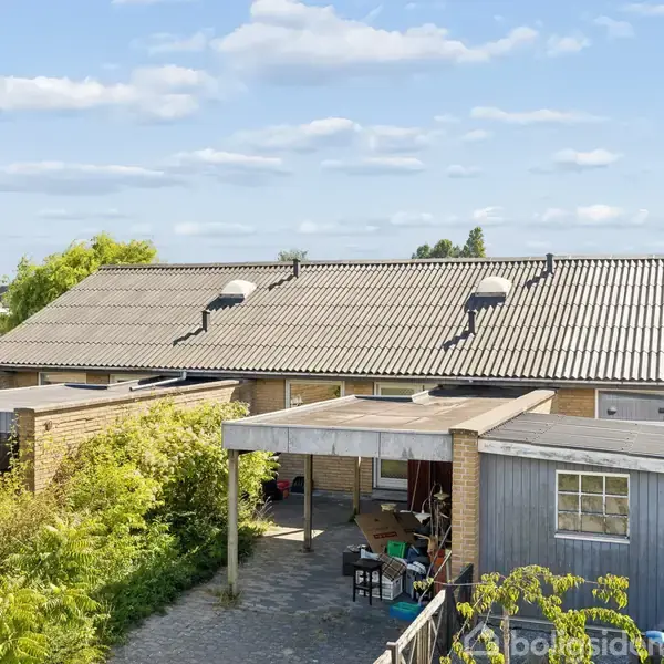 Rækkehus med fladt tag og carport, omkranset af grøn beplantning. Et spisebord og stole står under halvtaget. Biler er parkeret foran huset, mens himlen er skyet.