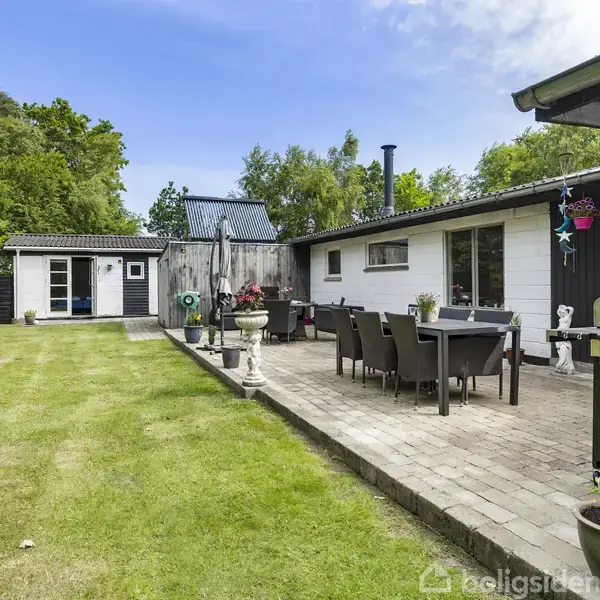 En terrasse med spisebord og stole står på fliser ved siden af et hus. En grill er placeret tæt på bordet. Græsplæne og træer omgiver området.