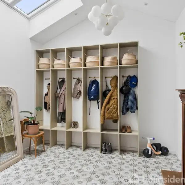 Garderobeskab fyldt med jakker, tasker og kasser er placeret i en entre med et mønstret flisegulv. Til venstre står et spejl og plante. Overhead hænger en moderne lampe.