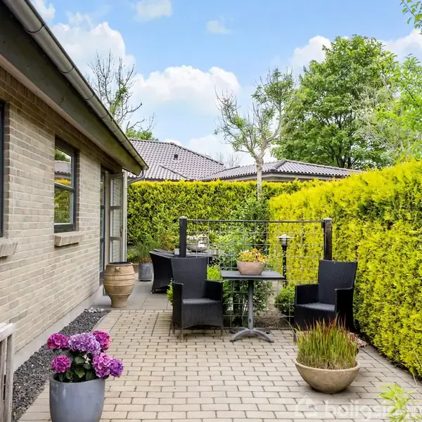 Terrassen har et lille bord med sort stolesæt omgivet af frodige planter og en høj hæk. De lyse murstenene på huset er synlige på venstre side.