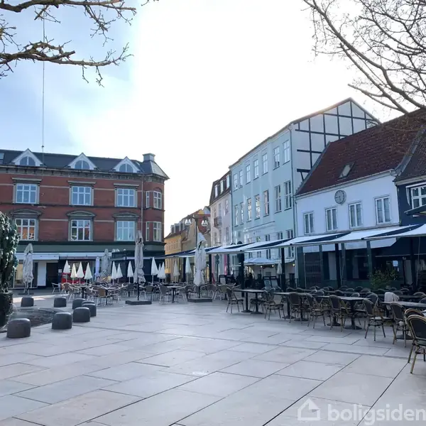 En række fortovscaféer står tomme på en brolagt plads omgivet af klassiske byhuse. Solen bryder gennem skydækket, og træer uden blade er synlige i hjørnerne.