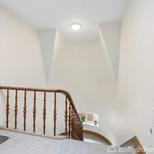 Trappeopgang med trægelænder. Trappen fører nedad i en hvid, minimalistisk korridor med et lille rundt vindue nederst, der lader lys trænge ind. En enkelt loftslampe oplyser rummet.