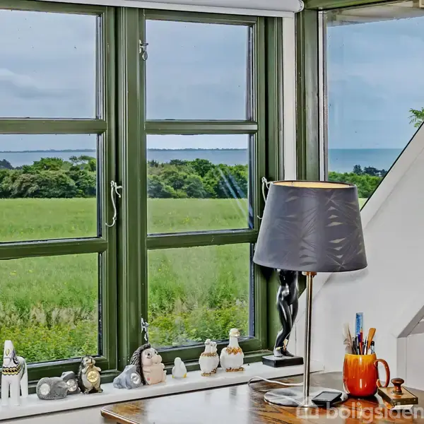 Et skrivebord med lampe står ved et vindue. Udsigt til grønne marker og havet i baggrunden. Små dekorationer og et indrammet foto findes på vindueskarmen.