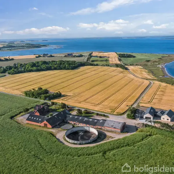 Gårdkompleks med flere bygninger ligger ved marker med høstede kornfelter. En vej snor sig gennem landskabet nær kysten, med udsigt over en bred blå fjord under en klar himmel.