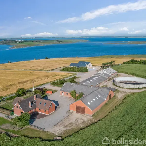 Gården består af røde murstensbygninger med gårdsplads og silo. Gården er omgivet af grønne marker og gyldne marker ved vandområde med blå himmel og skyer i baggrunden.