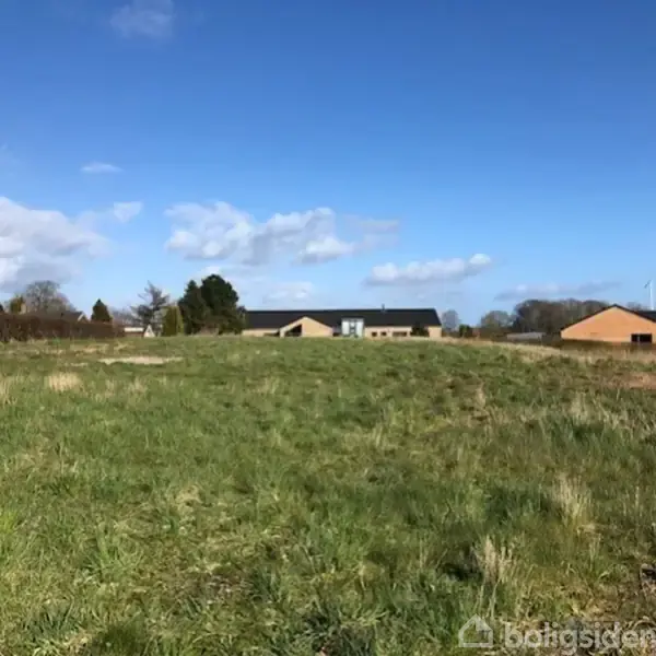 Grønt græsareal strækker sig mod flere huse i baggrunden under en blå himmel med få skyer.