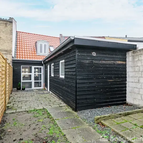 Sort skur står ved siden af brostensgang i en gårdhave med træ- og murstenshegn. Et rødt tegltag ses i baggrunden over et hus med to glasdøre.