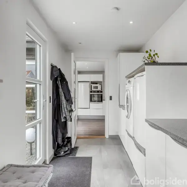Entré med vaskemaskine og tørretumbler placeret ved siden af en bænk. Jakker hænger på væggen. Døråbning fører til køkken med hvide skabe og ovn.