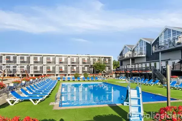 En swimmingpool med tilhørende liggestole omkring, omgivet af moderne bygninger og klart blå himmel, skaber en afslappende atmosfære i et ferieanlæg eller hotelkompleks.