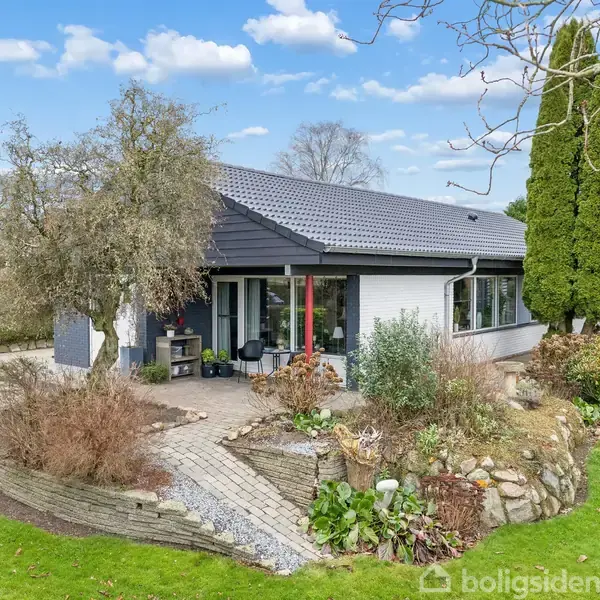 Et moderne hus står stille i en velplejet have med flisebelagt terrasse. Omgivet af træer og grønne buske under en blå himmel med få skyer.