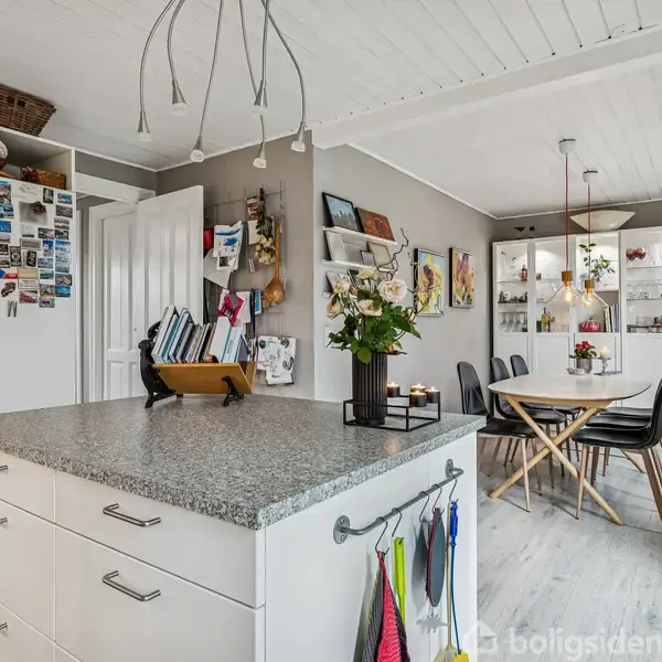 Køkkenø med grå bordplade har blomster i en vase, omgivet af køkkenskabe, malerier på væggen og et spisebord med sorte stole i et lyst rum.