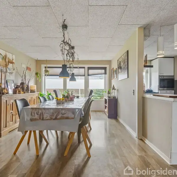 Spisebord med seks stole står i en lys spisestue. Bordet har en dug, og lamper hænger over det. Omgivet af planter og kunst på væggene i et moderne køkkenområde.