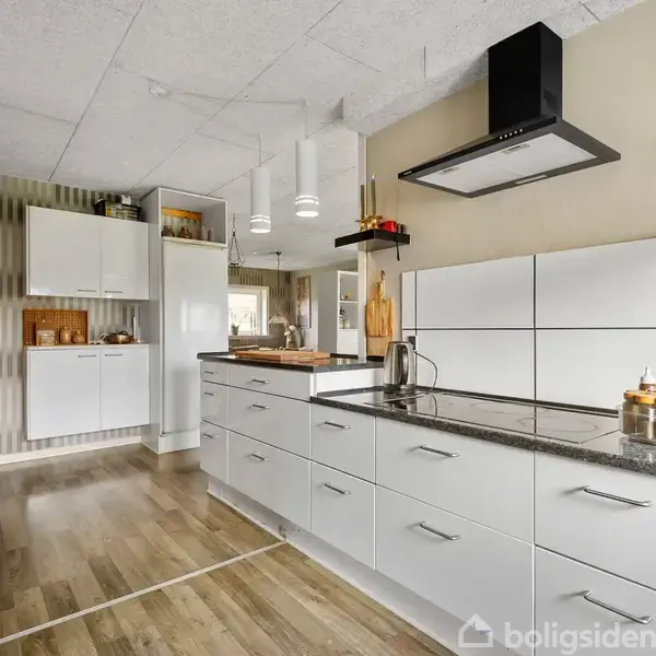 Moderne køkken med hvide skabe, sort bordplade og emhætte. Køkkenet er lyst med træ-laminatgulv og har indbyggede hylder og lysekroner, hvilket skaber en stilren og praktisk atmosfære.