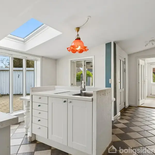 Et køkken med en hvid køkkenø er oplyst af en rød loftlampe. Store vinduer åbner ud mod en terrasse, og gulvet har et skakternet mønster.