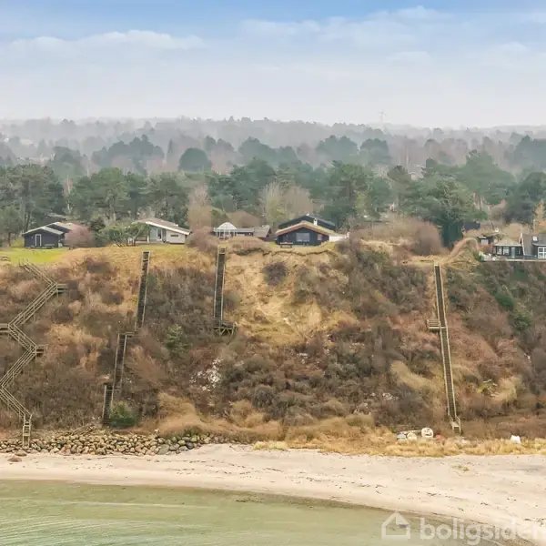 Flere sommerhuse er placeret på en høj klint med trapper ned til en sandstrand. Klinten er dækket af buske og træer, og baggrunden viser en skov.