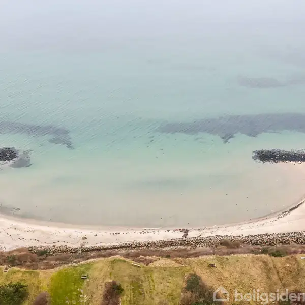 Strand med fint hvidt sand danner en lille bugt beskyttet af to bølgebrydere. Havet er roligt med grønt-blå vand. Forgrunden består af frodigt grønt terræn.