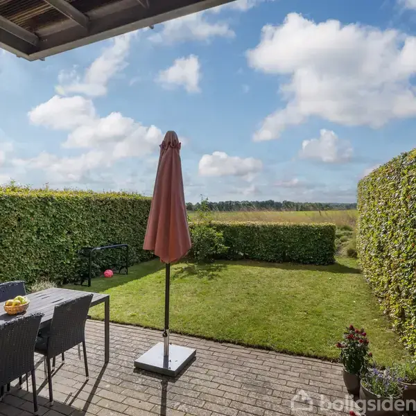 En terrasse med et bord, stole og en parasol står i en have omgivet af velplejede hække. Ud over hækkene ses en mark under en blå himmel med få skyer.
