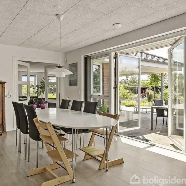 Et spisebord med sorte stole og to barnestole står i et lyst rum med trægulv. En skænk og en loftlampe ses, mens glasdøre åbner ud til en terrasse.