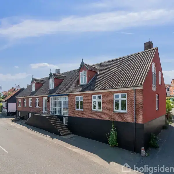 En stor, rød murstensbygning med skrånende tag står ved en vej. Bygningen har hvide vinduer og en trappe ved indgangen. Omgivet af andre huse og klar blå himmel.