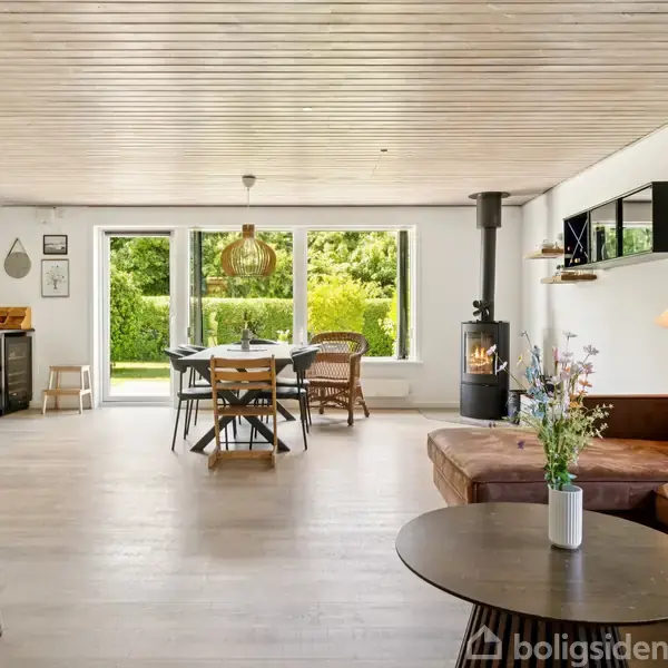 En lys stue med en brun lædersofa, sofabord med vase, og spisebord med stole ved vinduer. Brændeovn i hjørnet, hylder med dekorationer på væggen, og udsigt til have.