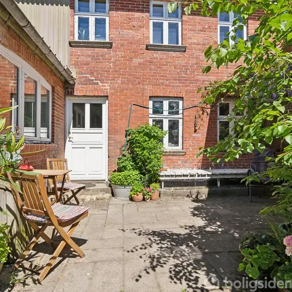 Stole og bord står på flisebelagt terrasse omgivet af grønne planter; rødt murstenshus i baggrunden med hvid dør og vinduer. Sollys skaber skygger på jorden.