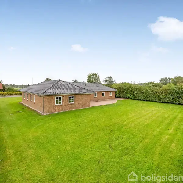 Et murstenshus står midt på en velplejet græsplæne omgivet af hække. I baggrunden er der træer og en rød bygning. Skyfri blå himmel danner en lys atmosfære.