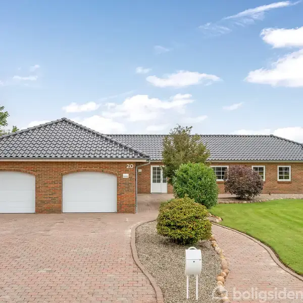 En murstensvilla med sort tegltag og dobbeltgarage står stille. Omgivet af velplejet græsplæne og buske. Husnummer "20" ses ved indkørslen. Klart vejr med blå himmel.