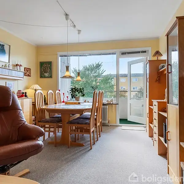 En stue med en brun lænestol står foran et spisebord med seks stole. Rummet er lyst med store vinduer og udsigt til en have. Reoler og lamper omkranser.