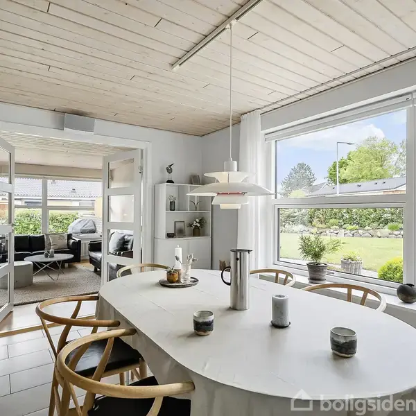 Et lyst spisebord med stole står i et spisestueområde, omgivet af store vinduer med udsigt til en have. En lampe hænger over bordet. I baggrunden ses en stue.