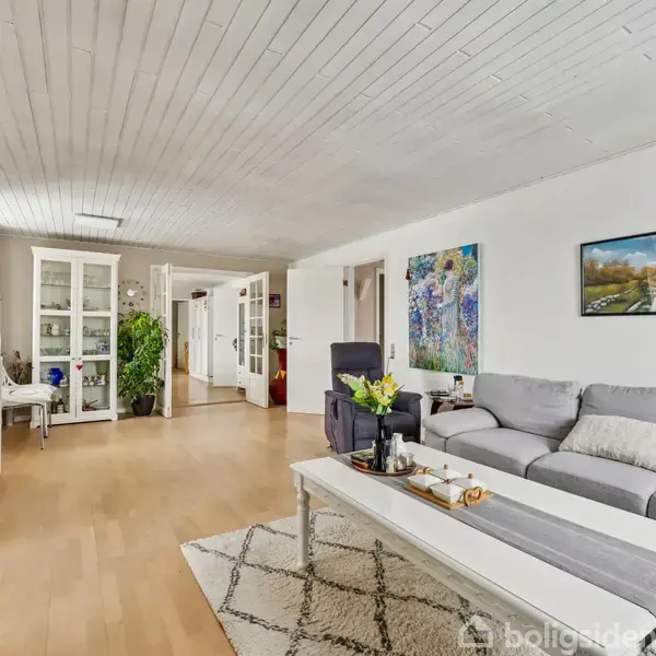 Stue med gråt hjørnesofa, sofabord med dekorationer, og tæppe. Væggene prydes af malerier. I baggrunden ses et skab, planter og en skrivebordstol. Lyst trægulv og hvidt loft.