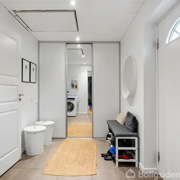 Entré med spejlvæg, sort bænk med puder, og to runde taburetter. Trægulvbelægning og lamper i loftet. Til højre en skydedør til et rum med vaskemaskine i baggrunden. Ingen tekst fundet.