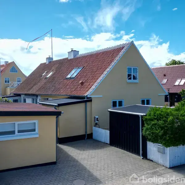 Gul, to-etagers hus med rødt tag står stille. Omkringliggende have med hæk og flisebelagt gårdsplads. Delft-blå himmel med skyer i baggrunden.