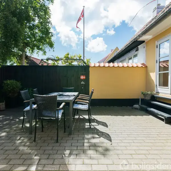 Terrasse med bord og stole står stille i sollys. Omgivet af gulmalet husvæg, sort hegn og planter. Dannebrog flagrer i baggrunden under blå himmel med skyer.