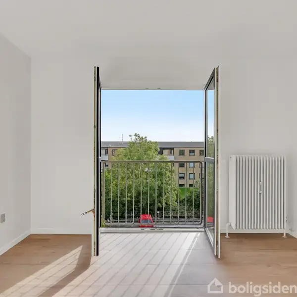 Dobbeltglasdøre i et hvidt rum åbner ud til en lille altan med udsigt til et boligområde og træer. På højre side er der en radiator.